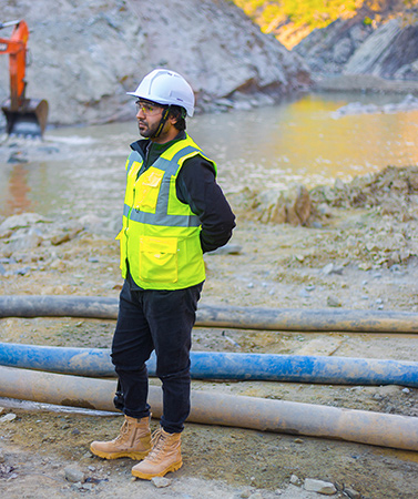 A worker in a hard hat and safety vest, at an outside work site.