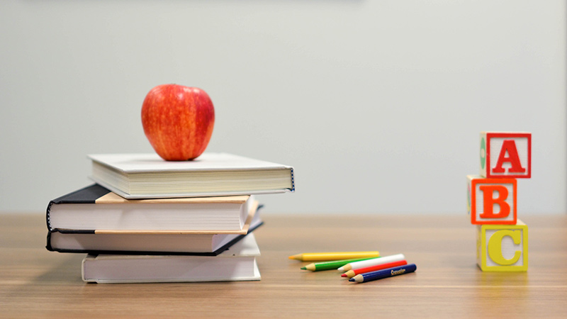 On the left a stack of 4 books with an apple on top. To the right 3 children's toy blocks, with colored pencils in between on the desktop.