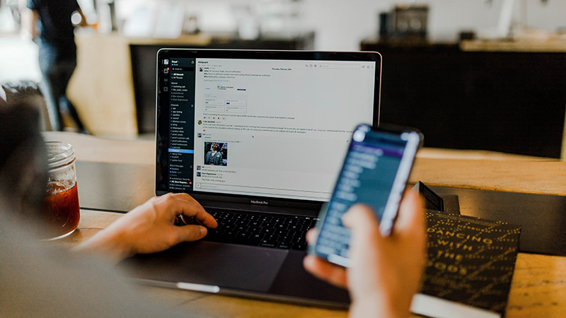 Open laptop on a desk with the user holding a phone in one hand.